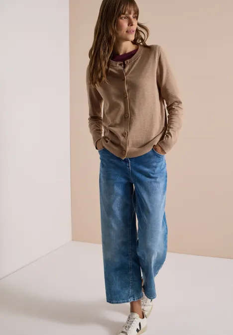 Vrouw leunt nonchalant tegen muur, draagt beige hoodie, wijde jeans en lichte sneakers.