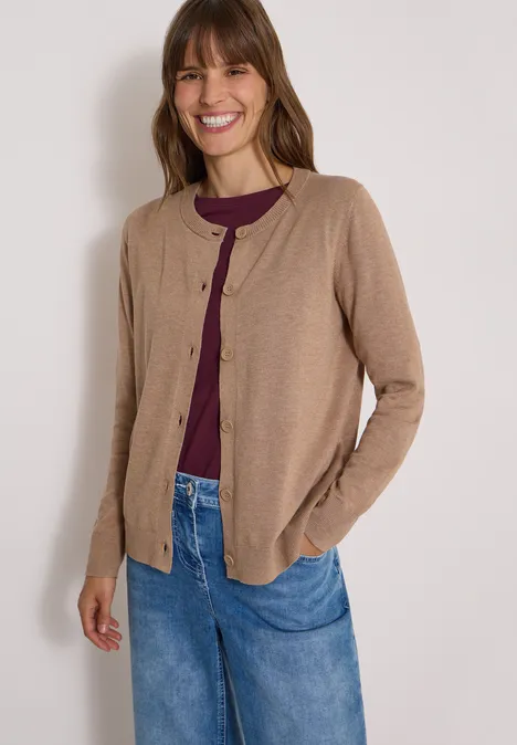 Een lachende vrouw draagt een beige vest over een paarse top en een spijkerbroek.