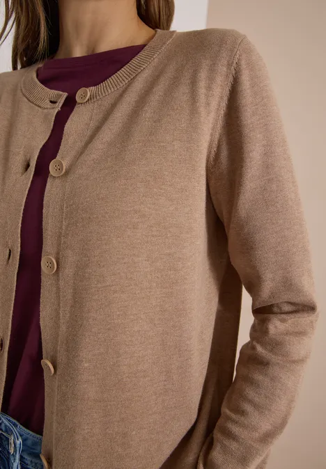 Vrouw in een beige gebreid vest met knopen over een donkerpaars shirt en blauwe jeans.