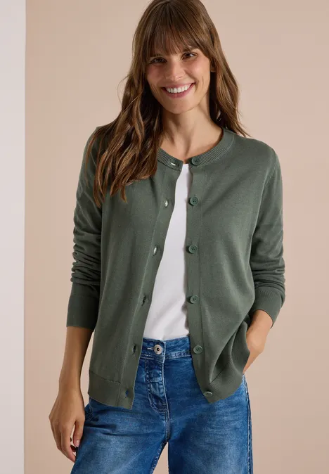 Lächelnde Frau in grünem Cardigan, weißem Shirt und Jeans vor neutralem Hintergrund.