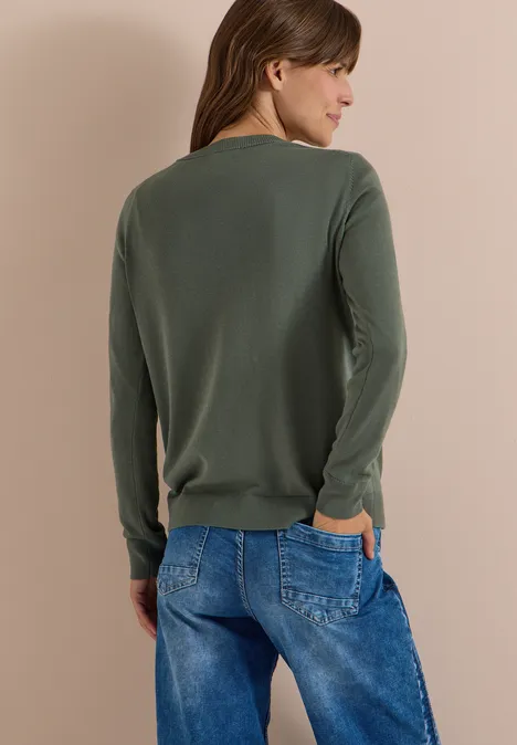 Eine Frau steht mit dem Rücken zur Kamera, trägt grünen Pullover und blaue Jeans vor hellem Hintergrund.