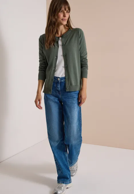 Eine Frau in grüner Strickjacke, weißem Shirt, blauen Jeans und Turnschuhen.