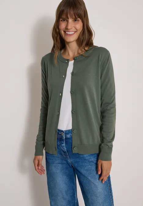 Frau lächelt in grüner Strickjacke, weißem Shirt und blauen Jeans vor weißem Hintergrund.