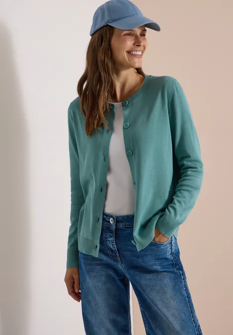 Eine lächelnde Frau trägt eine grüne Strickjacke, ein weißes T-Shirt, blaue Jeans und eine blaue Baseballkappe.