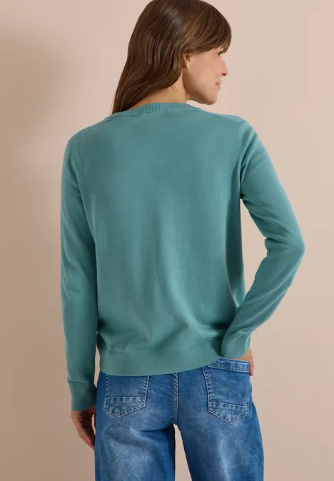 Frau von hinten in türkisfarbenem Pullover und Jeans vor neutralem Hintergrund.