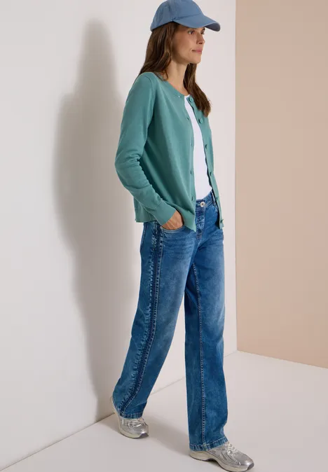 Frau lehnt an Wand, trägt grüne Strickjacke, weißes Shirt, weite Jeans, Sneaker und Cap.