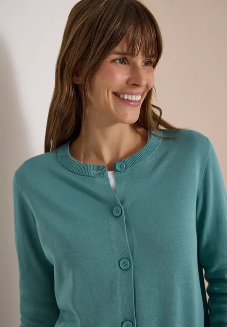 Frau in einem grün­türkisen Strickcardigan lächelt und blickt nach rechts.