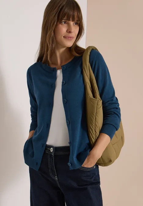 Cardigan à col rond et patte de boutonnage moonlight blue Eine junge Frau trägt eine blaue Strickjacke, weiße Bluse, Jeans und eine grüne Tasche.