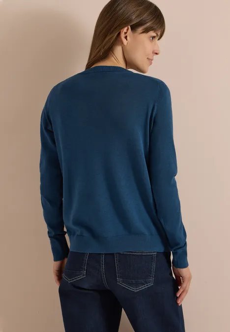 Cardigan à col rond et patte de boutonnage moonlight blue Eine Frau steht in dunkelblauem Pullover mit langen Ärmeln und Jeans, Aufnahme von hinten vor beigem Hintergrund.