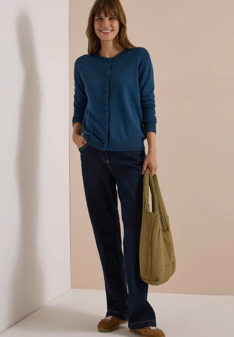 Cardigan à col rond et patte de boutonnage moonlight blue Frau in blauer Strickjacke und dunklen Jeans trägt eine grüne Tasche und lächelt vor schlichtem Hintergrund.