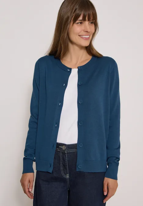 Cardigan à col rond et patte de boutonnage moonlight blue Frau mit blauer Strickjacke, weißem Shirt und Jeans lächelt seitlich.