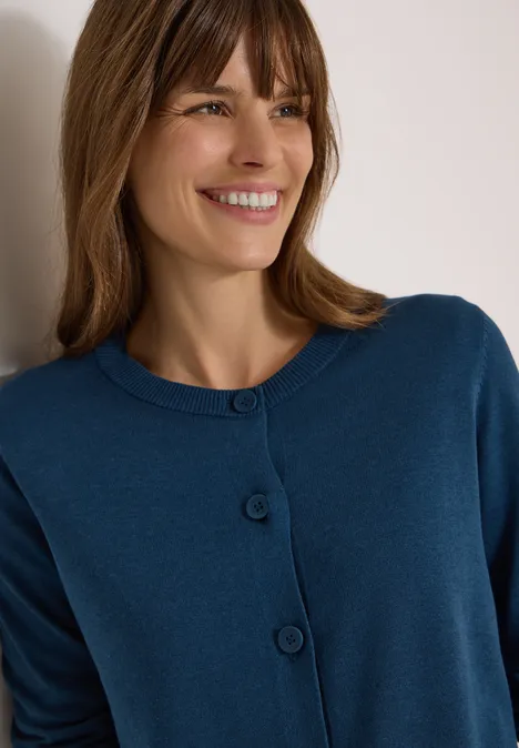 Cardigan à col rond et patte de boutonnage moonlight blue Frau lehnt lächelnd an Wand, trägt blauen Cardigan mit Knöpfen.