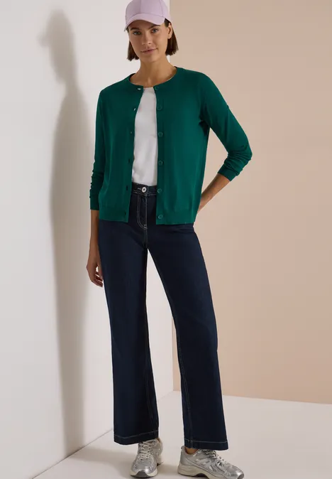 Cardigan boutonné à col rond dark fir green Femme en cardigan vert, t-shirt blanc, jean évasé bleu foncé et casquette rose.
