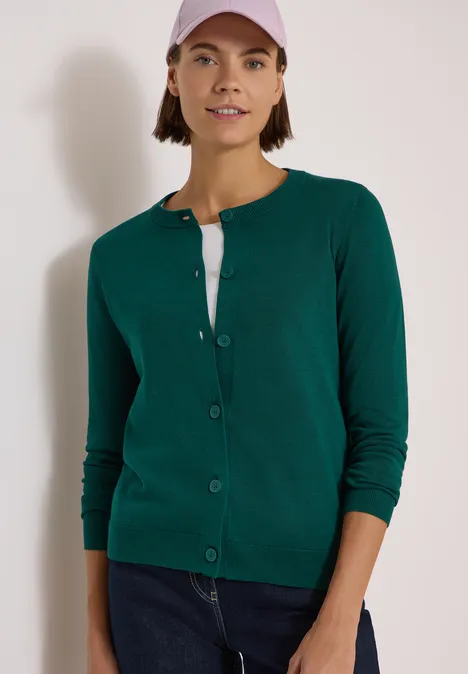 Cardigan boutonné à col rond dark fir green Femme en cardigan vert, t-shirt blanc, jean bleu et casquette rose devant un mur blanc.