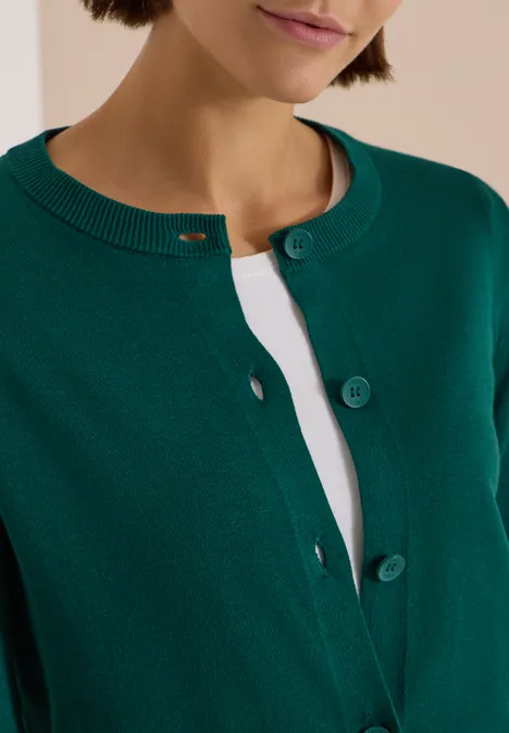 Cardigan boutonné à col rond dark fir green Cardigan vert à col rond et boutons, porté sur un haut blanc.