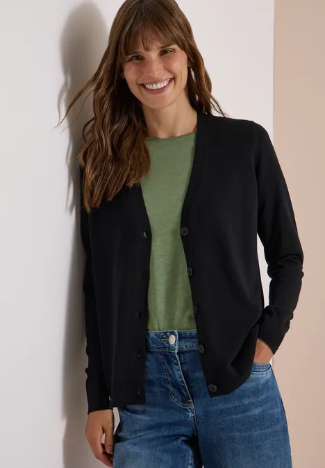 Vrouw glimlachend in jeans, groen t-shirt en zwarte cardigan bij een muur.