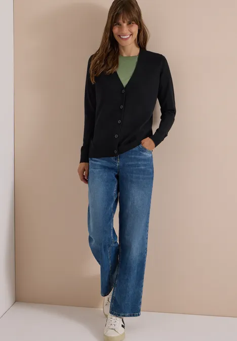 Een lachende vrouw draagt een zwart vest, een groen topje, wijd blauwe jeans en witte sneakers voor een beige muur.