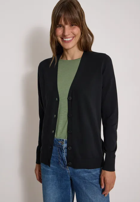 Een glimlachende vrouw draagt een zwarte cardigan, een groen shirt en blauwe spijkerbroek.