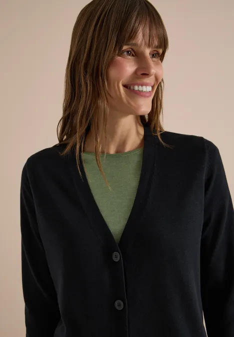 Een glimlachende vrouw met bruin haar draagt een zwart vest en een groen shirt.