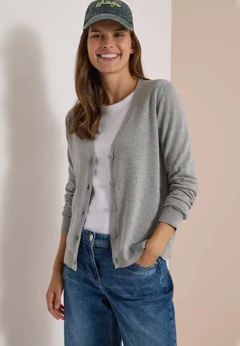 Cardigan boutonné à col V mineral grey melange Junge Frau mit grauem Cardigan, weißem T-Shirt, blauen Jeans und Baseballcap lächelt entspannt.