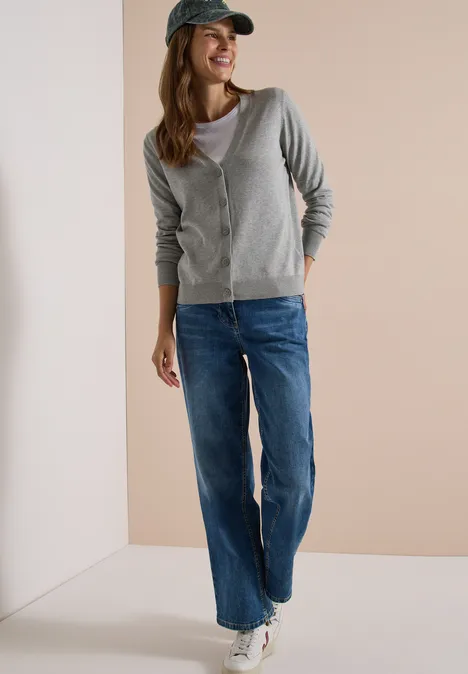 Cardigan boutonné à col V mineral grey melange Eine Frau in grauer Strickjacke, Jeans und Baseballkappe, lässig stehend mit Sneakern vor neutralem Hintergrund.