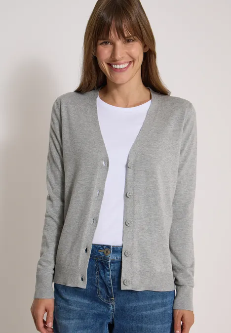 Cardigan boutonné à col V mineral grey melange Frau mit hellem Cardigan, weißem Shirt und Jeans lächelt vor hellem Hintergrund.