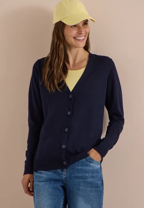 Vrouw lacht in donkerblauw vest, gele pet en spijkerbroek.