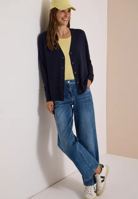 Jonge vrouw leunt tegen een muur, draagt geel topje, donkerblauwe cardigan, jeans, pet en witte sneakers.