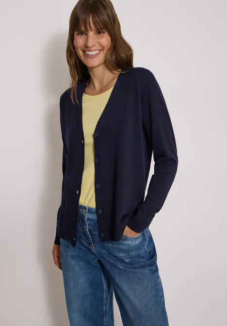 Vrouw lacht, draagt een donkerblauw vest, geel T-shirt en blauwe spijkerbroek.