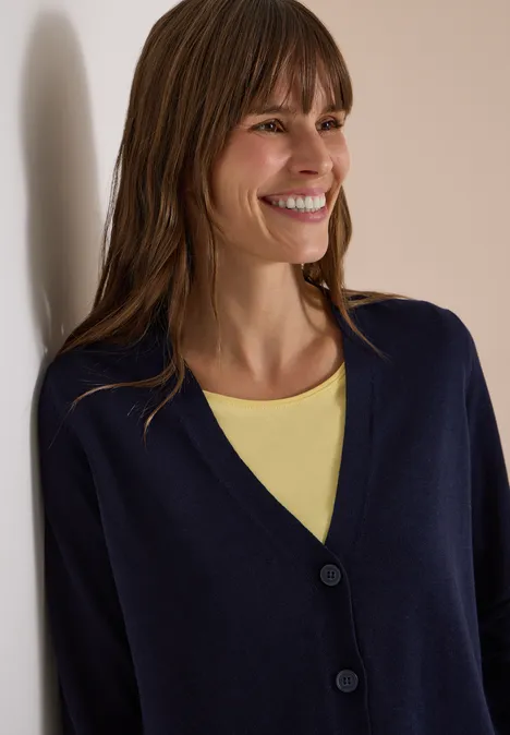 Lachende vrouw in marineblauw vest en geel topje leunt tegen een muur.