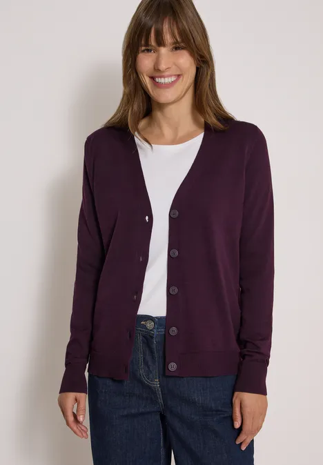 Kardigan z dekoltem w szpic i listwą guzikową eggplant red Uśmiechnięta kobieta w białym T-shircie, fioletowym kardiganie i dżinsach na jasnym tle.