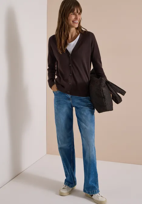 Vrouw in een casual vest, spijkerbroek en sneakers, houdt een jas vast tegen neutrale achtergrond.