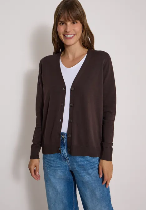 Vrouw glimlacht in een bruine cardigan, wit shirt en spijkerbroek, voor een neutrale achtergrond.