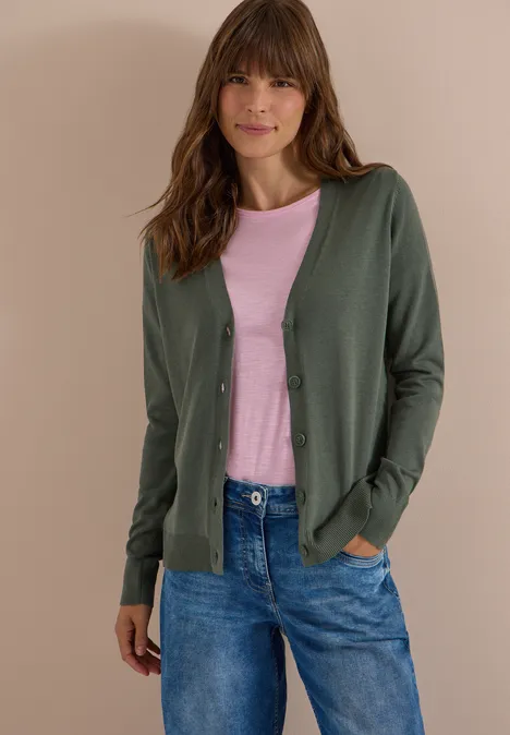 Cardigan mit V-Neck und Knopfleiste dried olive green Frau trägt grünen Cardigan, rosa Shirt und Jeans, steht vor neutralem Hintergrund mit Händen in Taschen.