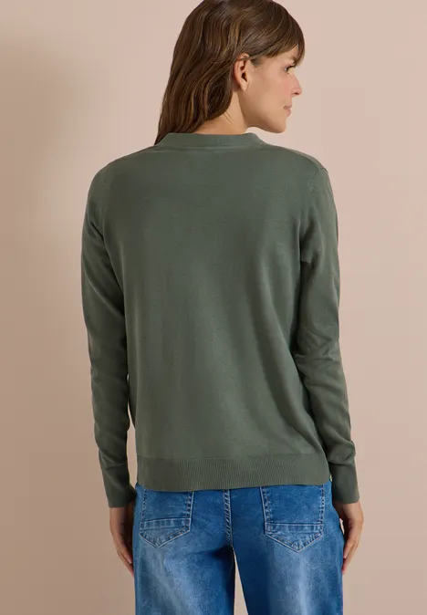 Cardigan mit V-Neck und Knopfleiste dried olive green Eine Frau steht seitlich gewandt vor beigem Hintergrund, trägt einen grünen Pullover und blaue Jeans.