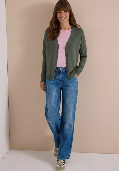 Cardigan mit V-Neck und Knopfleiste dried olive green Frau in grüner Strickjacke, rosa T-Shirt und weitem Jeansbund posiert vor beiger Wand.