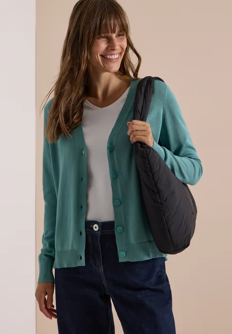 Vrouw lachtend in een turquoise cardigan met wit shirt, donkerblauwe jeans en zwarte schoudertas.