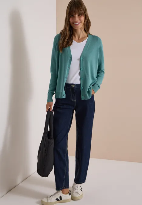Een lachende vrouw in een groen vest, wit topje, donkere broek en sneakers, met een zwarte tas voor beige achtergrond.