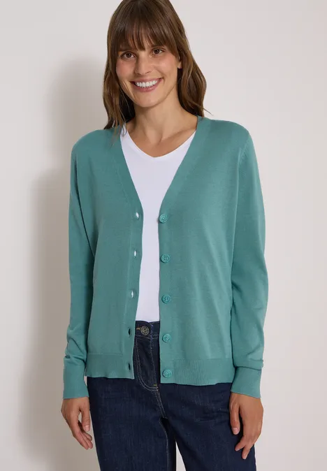 Vrouw met groene cardigan, wit T-shirt en donkere jeans, glimlachend voor een neutrale achtergrond.