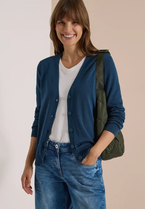 Cardigan boutonné à col V moonlight blue Frau in weißem T-Shirt, blauem Cardigan und Jeans trägt grüne Tasche und lächelt.