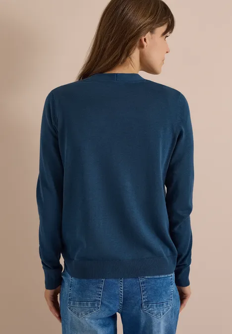 Cardigan boutonné à col V moonlight blue Frau von hinten in blauem Pullover und Jeans vor beigem Hintergrund