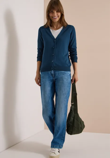 Cardigan boutonné à col V moonlight blue Frau in blauem Cardigan, Jeans und weißem Shirt hält grüne Tasche vor hellem Hintergrund.