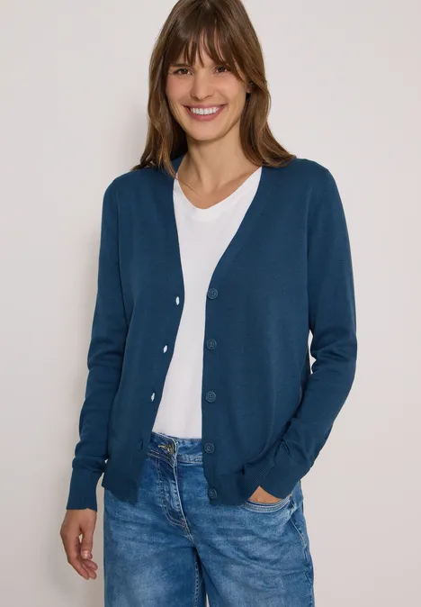 Cardigan boutonné à col V moonlight blue Eine Frau trägt einen blauen Cardigan, weiße Bluse und Jeans, lächelt vor hellem Hintergrund.