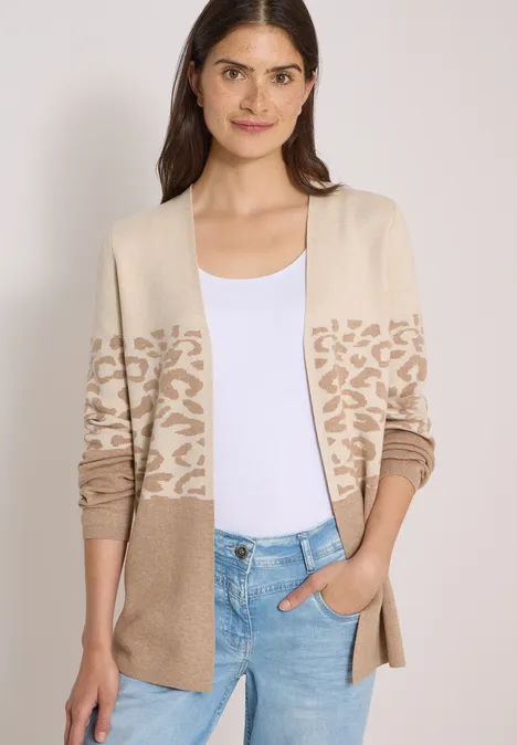 Eine Frau trägt einen beigen Cardigan mit Muster über weißem Top und blauer Jeans.
