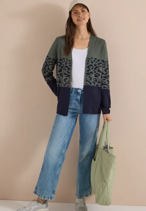 Frau im Cardigan mit grün-schwarzem Muster, weißem Shirt, lockeren Jeans und grüner Tasche.