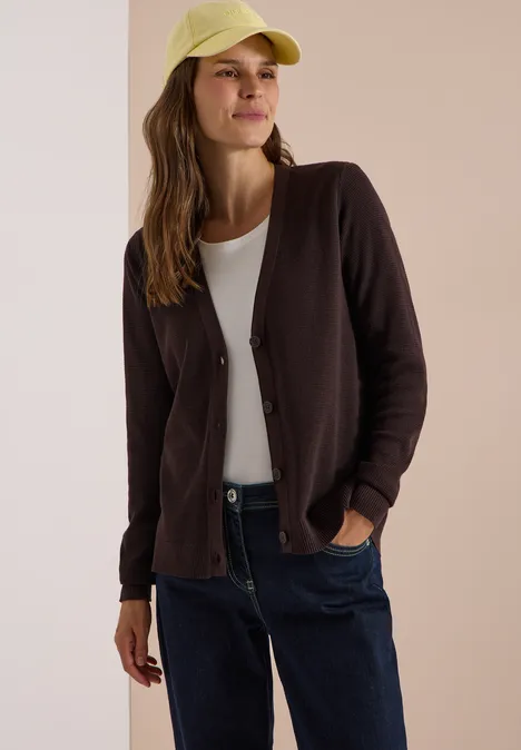 Kardigan z dekoltem w szpicu i strukturą truffle brown Uśmiechnięta kobieta w brązowym kardiganie, białym T-shircie, niebieskich dżinsach i żółtej czapce.