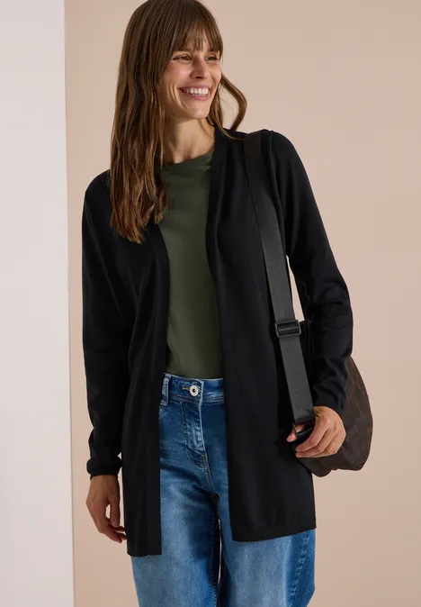 Een vrouw draagt een zwarte cardigan, groen shirt, jeans en lacht, met een tas over de schouder.