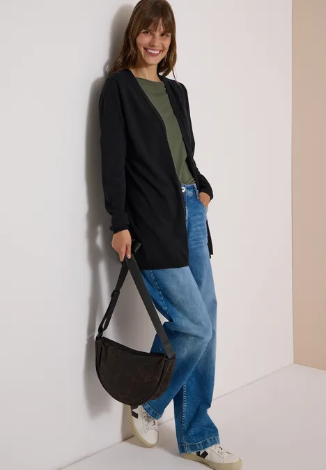 Vrouw leunt tegen muur, draagt zwarte cardigan, groen shirt, wijde spijkerbroek, witte sneakers en zwarte tas.