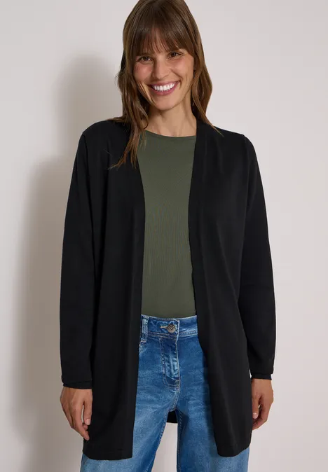 Vriendelijke vrouw in zwart vest, olijfgroen shirt en blauwe jeans tegen een neutrale achtergrond.