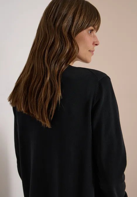 Vrouw gezien van achteren met golvend bruin haar, zwarte trui, lichte achtergrond, hoofd licht gedraaid.